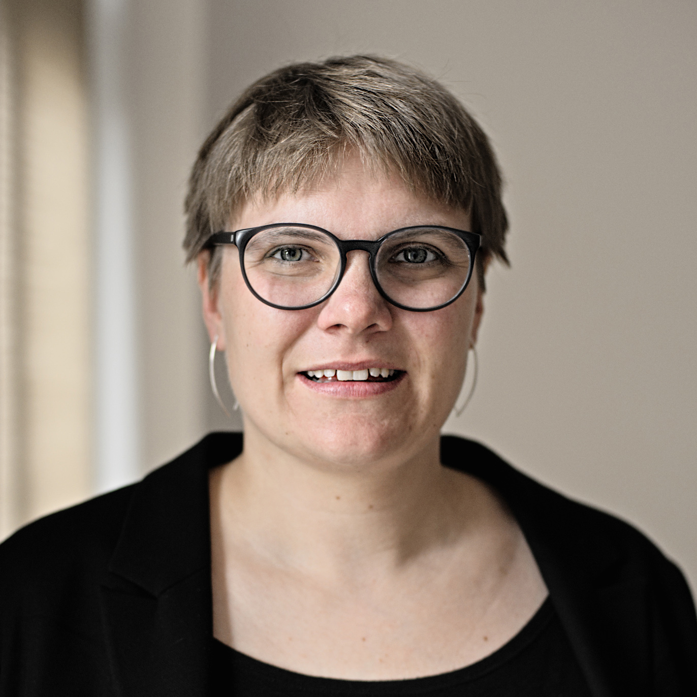 Portrait von Luise Veit. Sie lacht freundlich nach vorn. Luise Veit trägt kurze Haare, eine Brille mit dunklem Rand und zwei silberne Ohrringe. Sie trägt ein schwarzes Oberteil und einen schwarzen Blazer. Im Hintergrund ist eine helle Wand.
